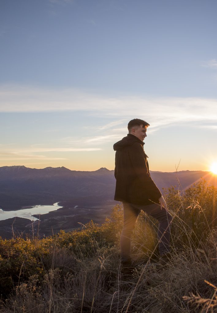 A white man stands on a golden cliff in front of a purple mountain range, watching the sun rise into the bright blue sky.