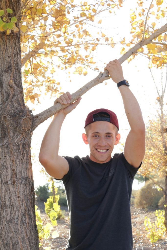 A latine man wearing a grey shirt and a red backwards snapback stands against a fall foliage background. He is grinning and reaching up to clasp the tree branch above him.