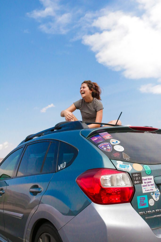 a latine girl with brown hair sits atop a blue subaru that has stickers covering the back. There is a blue sky behind her with white clouds floating in the same direction as the car is facing.