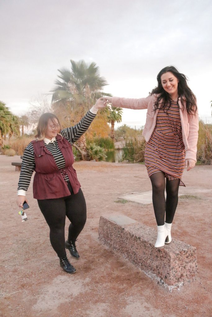 A girl in a black stripped top and red vest holds hands with a brown haired girl wearing a pink stripped dress and fluffy pink jacket, as the laugh and walk towards the camera.